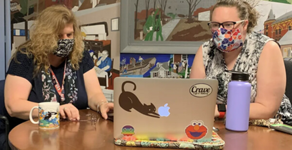 Photo of two ladies in masks at computer on table perform peer specialist duties