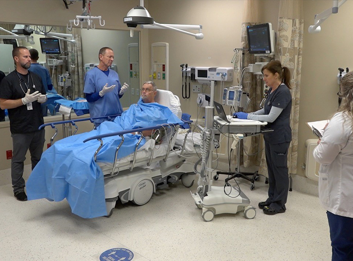 Four simulation experts meeting around a high fidelity manikin in a hospital room.